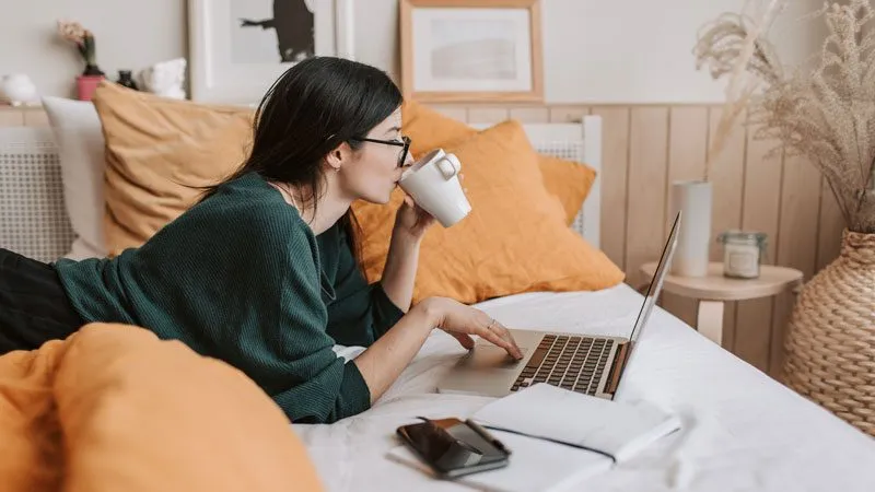 Frau mit Kaffeetasse liegt im Bett und tippt auf einem Laptop