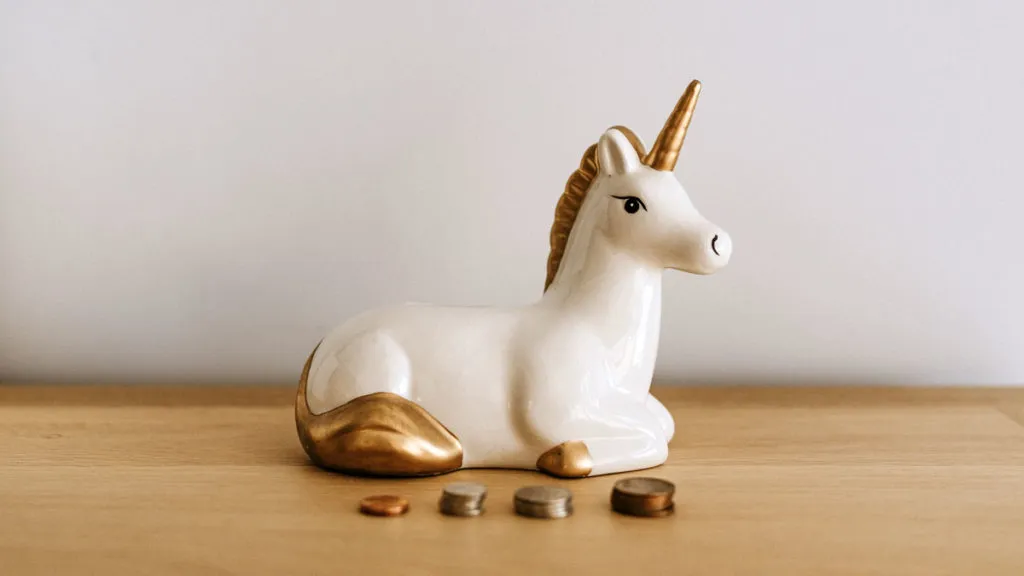 Weiß-goldene Einhorn-Spardose auf einem Holztisch, davor vier kleine Münzstapel