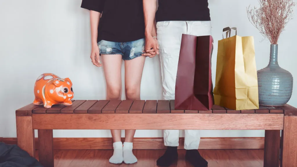Zwei Menschen halten Händchen hinter einer Holzbank, links eine orange Sparschwein-Figur, rechts zwei Einkaufstüten